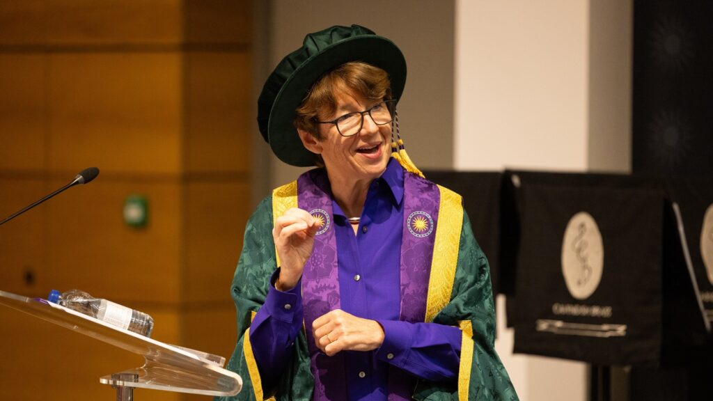 Chancellor Dawn Airey wears her cap and gown and stands next to a lectern while looking towards the graduates.
