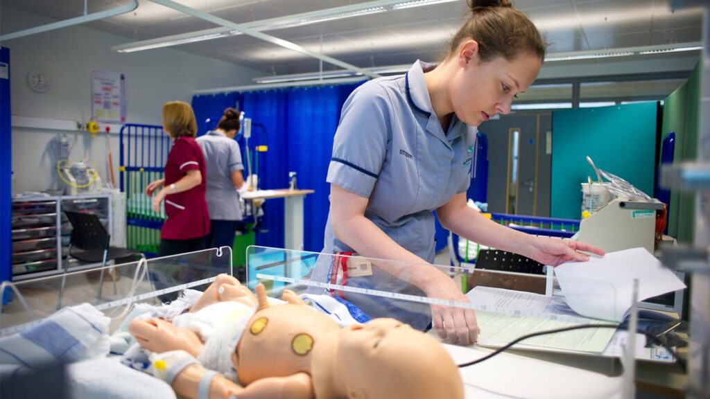 A student nurse standing over a doll and reading its notes.
