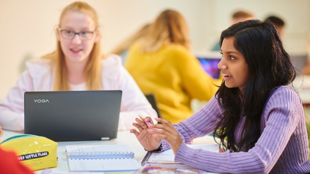Two students talking whilst at a classroom table. They have their notepads and one student has their laptop.