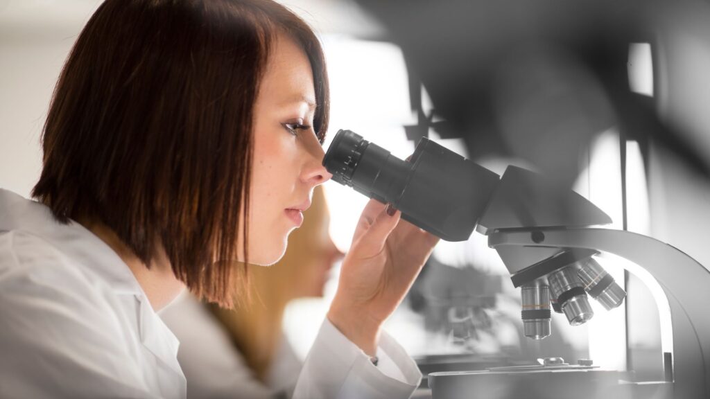 A student in a lab coat looks down the lens of a microscope.